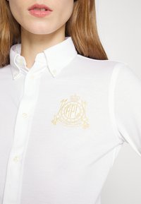 Vit polo-tröja med knapp-down krage. Har en guld broderad logotyp på bröstet. Texturerat tyg med figursydd design.