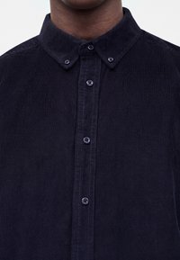 Homme portant une chemise en velours côtelé bleu foncé à col boutonné, montrée du cou jusqu'au milieu de la poitrine.