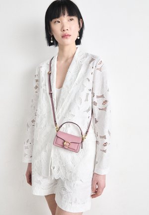 Kvinde med kort sort hår iført hvid blazer og shorts i blomsteragtet blonde, accessoriseret med perleøreringe og en lille pink crossbodytaske.