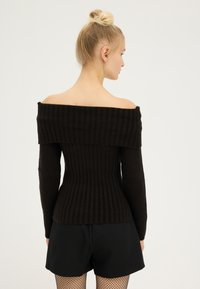 Crni pulover s off-shoulder dizajnom od rebrastog pletenog materijala, dugih rukava, s uskim krojem i teksturiranim završetkom. Uparen s crnim šorcem.