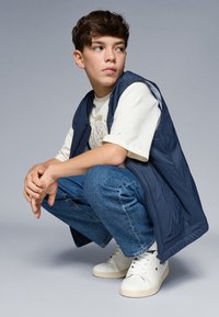 Gilet senza maniche navy sopra una maglietta chiara, abbinato a jeans blu e sneakers bianche. Tessuto strutturato con dettagli a zip.