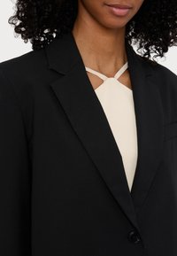 Blazer noir avec un col cranté, fermeture à un bouton, et une poche, porté sur un haut beige clair avec un détail découpé.