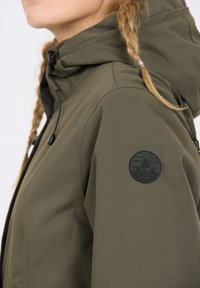 Olivgrön jacka med texturerat ytskikt, försedd med en svart rund lapp på axeln och justerbara dragsnören på huvan.