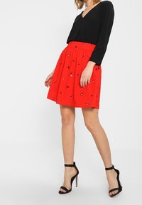 Haut noir à manches longues avec un col en V, associé à une jupe rouge à taille froncée et motifs brodés. Sandales noires à talons hauts.