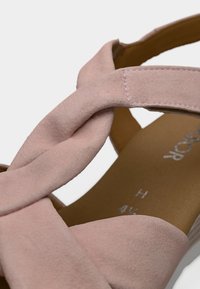 Ροζ σανδάλι από σουέντ με σχεδιασμό στριφτού λουριού, καφέ πάτο και μινιμαλιστικές λεπτομέρειες ραφής. Στυλ με ανοικτή μύτη και λουρί στον αστράγαλο.