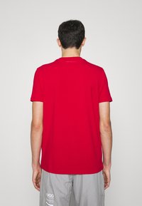 Červené tričko s krátkým rukávem, které má kulatý výstřih a hladkou texturu. Logo značky je decentně tištěno na zadní straně blízko výstřihu.