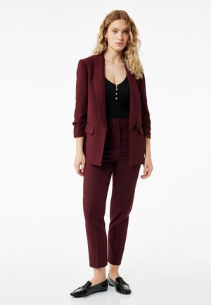 Kobieta o długich blond włosach, ubrana w bordowy garnitur z czarnym topem i czarnymi mokasynami, stojąca na białym tle.