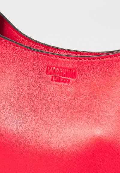 Punainen nahkalaukku, jossa on sileä pinta. Varustettu kohotetulla "Moschino Milano" -logolla, jossa on musta reunus ylhäällä.