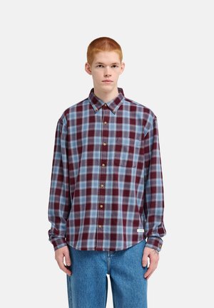 Young man with short red hair wearing a loose blue and burgundy checkered button-up shirt and blue jeans, standing against a plain background.