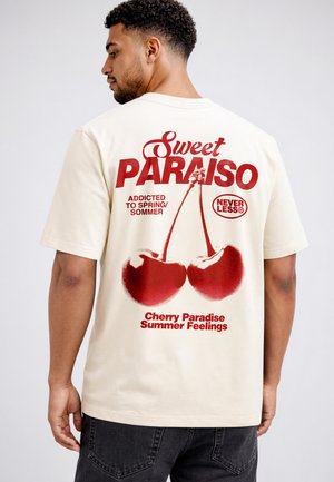 Mann mit kurzem lockigem Haar, der ein cremefarbenes T-Shirt mit rotem Text und einer Grafik von zwei Kirschen auf dem Rücken trägt, seitlich nach links blickend.