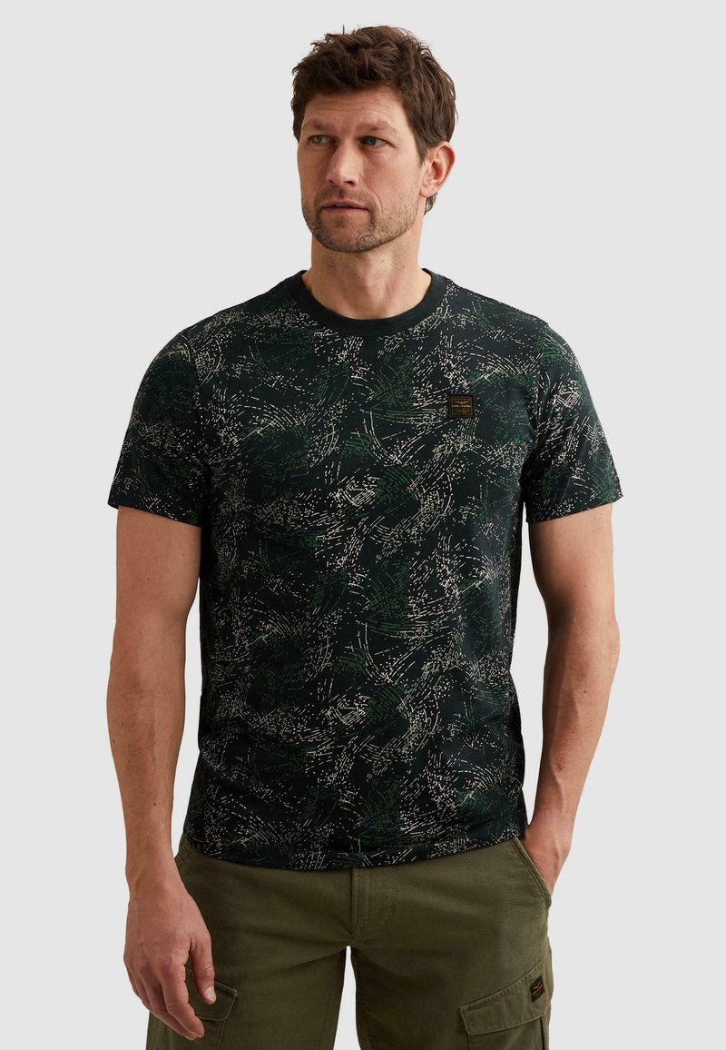 Mann trägt ein schwarz-grünes, abstrakt gemustertes T-Shirt mit kurzen Ärmeln und olivgrüne Cargohosen vor neutralem Hintergrund.