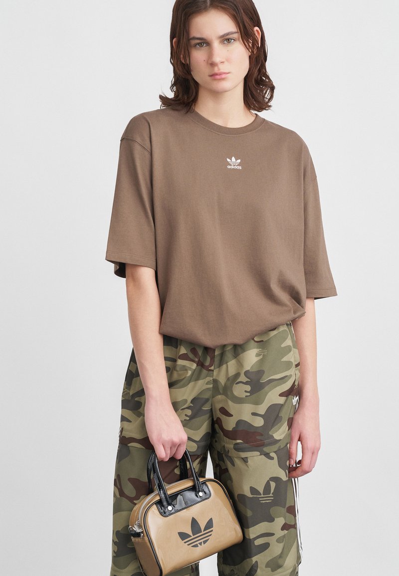 Tricou maro oversized cu logo-ul alb Adidas, asortat cu pantaloni verzi cu camuflaj. Într-o mână este ținută o geantă mică bej cu accente negre.