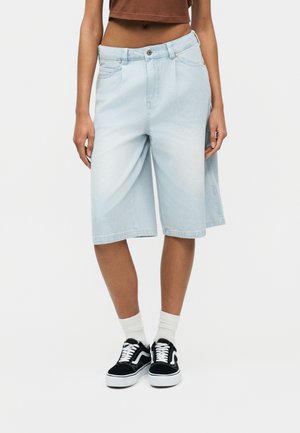 Άτομο που φοράει γαλάζια τζιν σορτς μέχρι το γόνατο, λευκές κάλτσες και μαύρα και άσπρα αθλητικά παπούτσια, στέκεται σε απλό φόντο.