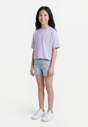 Lány áll és mosolyog, világoslila rövid ujjú inget, farmer rövidnadrágot és fehér tornacipőt visel, fehér háttér előtt.