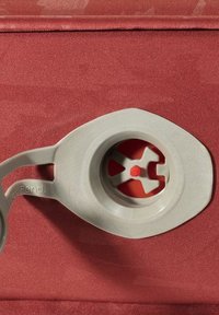 Grijze plastic klep met een intern rood mechanisme, bevestigd aan een gestructureerde rode stoffen basis. De klep is ovaalvormig met een verhoogde rand.