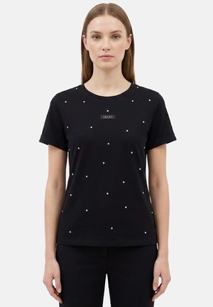 Donna con capelli lisci castano chiaro indossa una maglietta nera a maniche corte con piccoli borchietti argentati e pantaloni neri, in piedi su sfondo bianco.