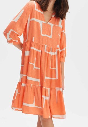 Donna che indossa un abito arancione largo fino al ginocchio con motivi geometrici bianchi a quadrati e maniche a sbuffo a tre quarti.