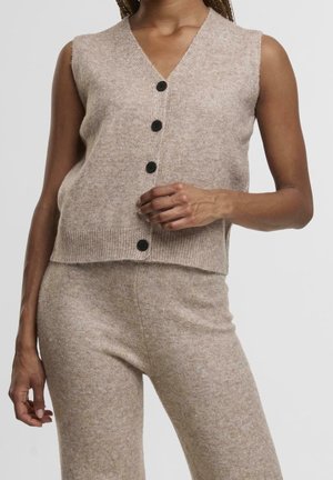 Frau trägt eine beige ärmellose Strickweste mit Knöpfen und passende, hoch taillierte Strickhosen vor einem schlichten Hintergrund.