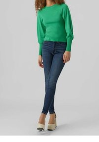 Vihreä neulepusero, jossa on pyöreä kaula-aukko ja puffi hihnat, yhdistettynä slim fit -tummansinisiin farkkuihin ja kermaiset korkokengät.