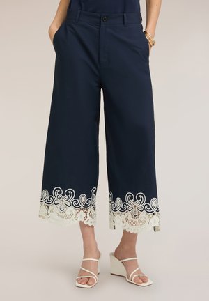 Marineblauwe wijde cropped broek met witte kanten bloemendetail aan de zoom, gedragen met witte open-toe sleehakken, handen in de zakken.