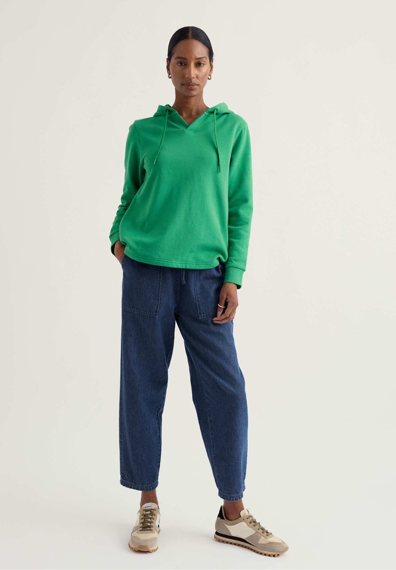 Frau trägt einen leuchtend grünen Kapuzenpullover, weite dunkelblaue Jeans und beige Turnschuhe, steht vor einem schlichten hellen Hintergrund.