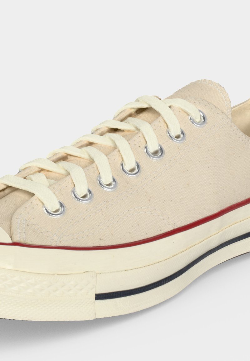 Sneaker beige in tela bassa con lacci bianchi, occhielli in metallo e suola spessa con strisce rosse e nere.