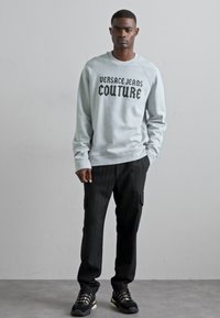 Světlemodrá mikina se nápisem "Versace Jeans Couture" v černém písmu, kombinovaná s černými pruhovanými kalhotami a černými teniskami.