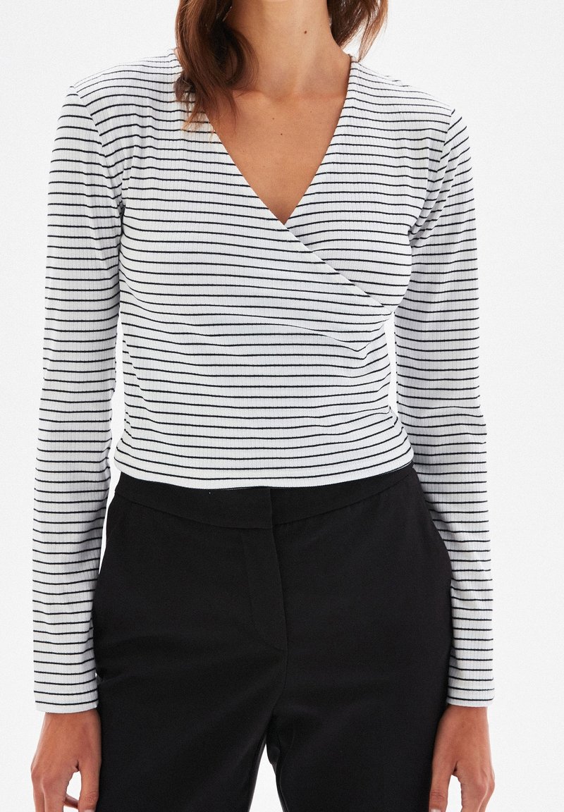 Femme portant un haut blanc à manches longues avec de fines rayures noires horizontales et un pantalon noir, torse visible sur un fond blanc.