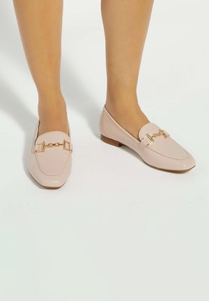 Mocassins en cuir rose pâle avec détail de chaîne dorée, portés pieds nus, posés sur une surface blanche.