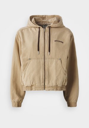 Beige hettejakke laget av bomull med glidelås foran, elastiske mansjetter og liv, med to frontlommer og brodert merke.