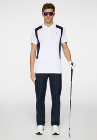 Vit polo med marinblå detaljer, kortärmad; i kombination med marinblå cargobyxor. Bär glasögon och håller en golfklubba, med vit golfhandske.