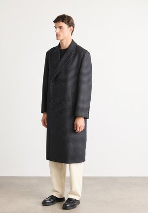 Young man standing wearing a long dark grey houndstooth double-breasted coat, cream trousers, and black loafers on a plain background.