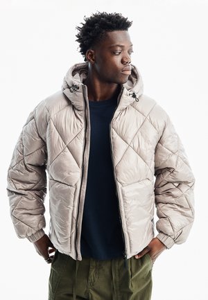 Junger Mann trägt eine hellbeige gesteppte Kapuzenjacke über einem schwarzen Hemd und grünen Hosen, schaut zur Seite mit den Händen in den Taschen.