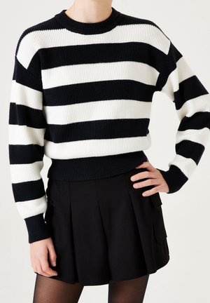 Personne portant un pull à rayures horizontales noires et blanches avec une jupe plissée noire et des collants noirs transparents.