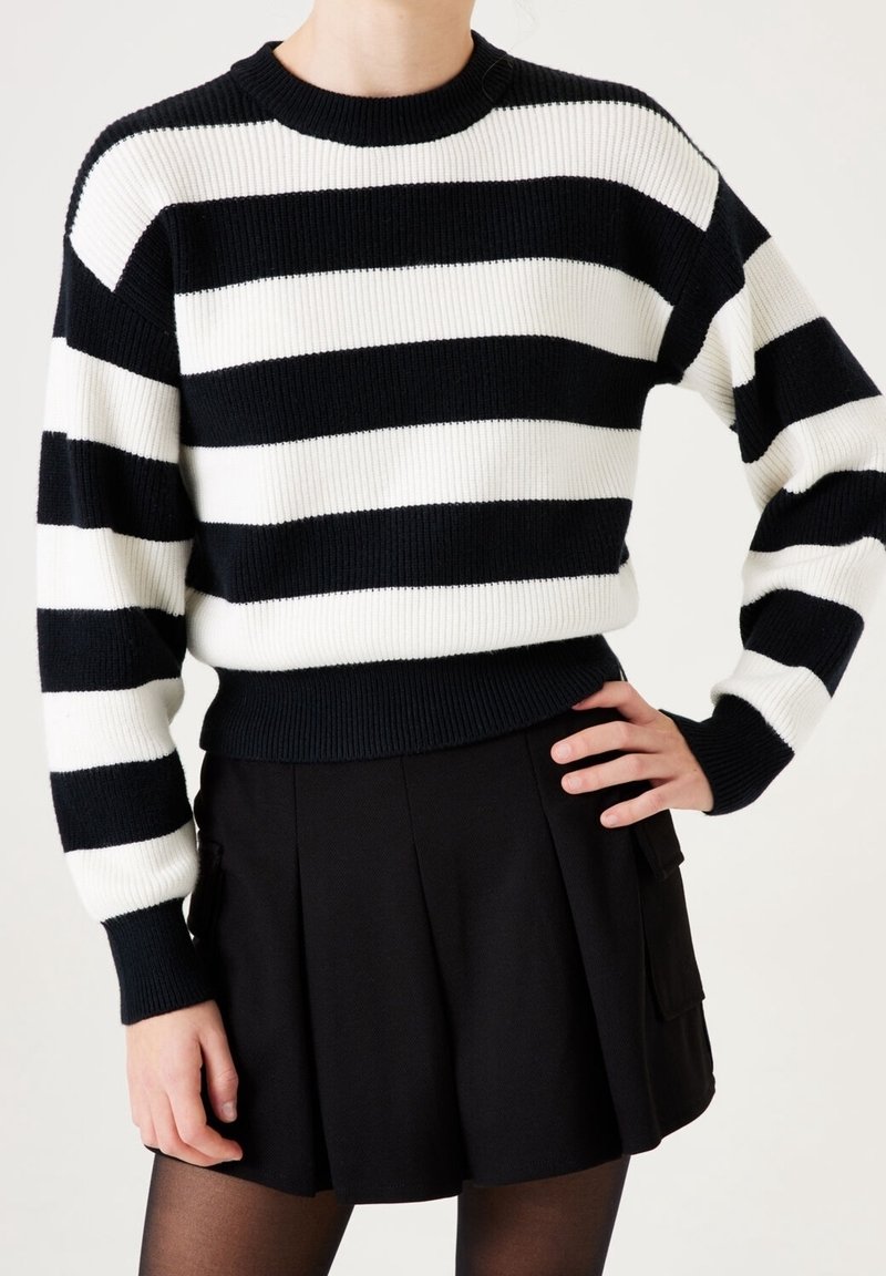 Personne portant un pull à rayures horizontales noires et blanches avec une jupe plissée noire et des collants noirs transparents.
