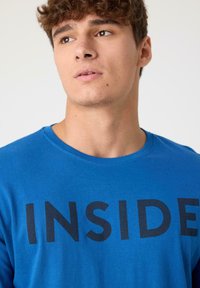 Camiseta de algodón azul con cuello redondo que presenta la palabra "INSIDE" en letras negras en negrita a lo largo del pecho. Textura suave.