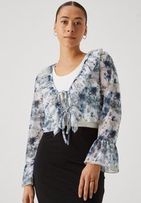 Blus med blommönster i genomskinligt material, med volangdetaljer vid halsen och ärmsluten, huvudsakligen i blått och vitt, med en knytning fram.