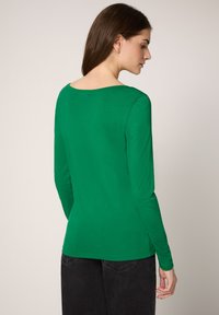 Vihreä, pitkähihainen, istuva paita kirkkaasta kankaasta. Siinä on leveä kaula-aukko ja sileä pinta, yhdistetty tummiin farkkuihin.