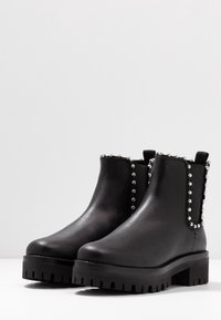 Botas de cuero negras hasta el tobillo con suela gruesa, que presentan tachuelas plateadas a lo largo del costado y paneles elásticos para facilitar su uso.