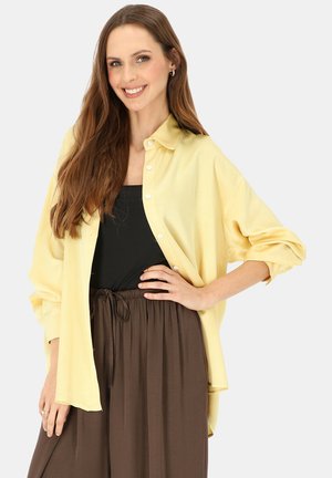 Kobieta uśmiechająca się z długimi brązowymi włosami, ubrana w luźną żółtą koszulę na guziki na czarny top i brązowe spodnie ze sznurkiem.