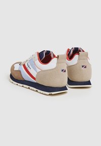 Pár béžových, bílých, červených a modrých volnočasových tenisek s semišovými detaily, červeným podkladem a logovými nášivkami na patě a bočních panelech.