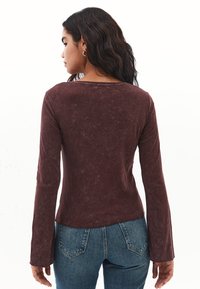 Langärmliges, kastanienbraunes Oberteil mit weicher, strukturierter Oberfläche. Verfügt über einen runden Halsausschnitt und ausladende Ärmel, kombiniert mit blauen Jeans.