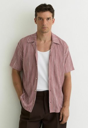 Man met een openhangend roze-wit gestreept overhemd met korte mouwen over een wit hemd, staand tegen een witte muur.