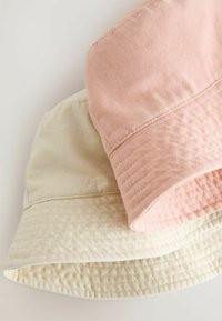 Två bucket hats visas, en rosa och en beige, gjorda av texturerad bomull med en mjuk, flexibel brätte och sydda detaljer.