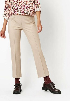 Woman wearing beige straight-leg trousers, floral long-sleeve blouse, maroon socks, and dark brown lace-up shoes, standing against white background.