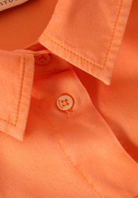 Orange collared shirt with smooth texture, two buttons featuring a flower design, and neat stitching along collar edges.