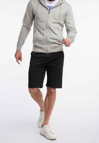 Felpa grigia con zip e lacci, indossata sopra una maglietta bianca, abbinata a pantaloni corti neri e sneaker bianche, che mostrano un outfit casual.