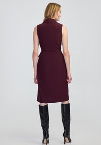 Donna che indossa un abito borgogna senza maniche, lungo fino al ginocchio, e stivali neri con tacchi alti fino al ginocchio, in piedi di spalle su uno sfondo semplice.