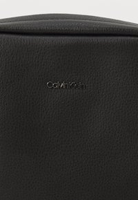 Černá kožená taška Calvin Klein s texturovaným povrchem. Vyznačuje se elegantním designem a výrazným stříbrným logem na přední straně.