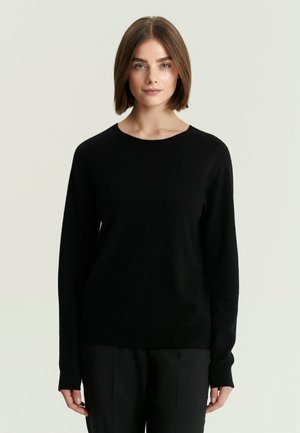 Mujer con cabello corto y castaño que lleva un jersey negro liso de manga larga y pantalones negros, de pie frente a un fondo claro y neutro.